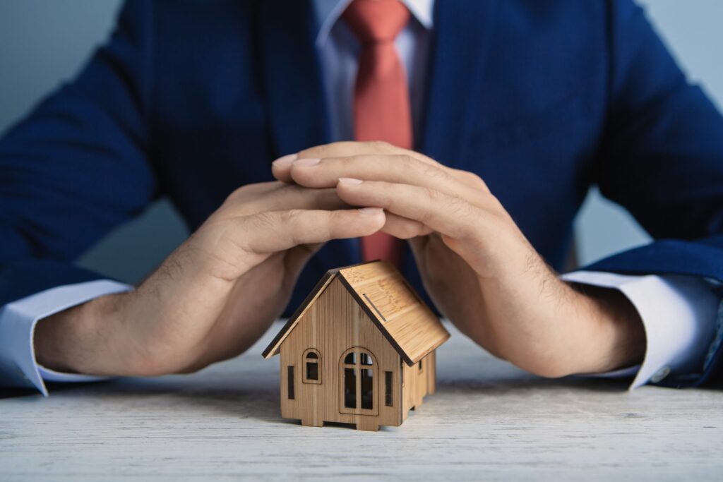 A man in a suit protects a small wooden houses with his hands, representing someone preventing home security problems.